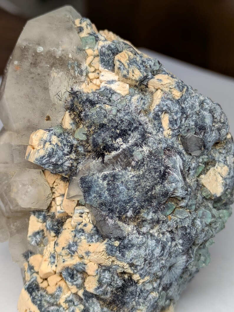 May include: A close-up of a natural mineral specimen. The crystal formation features a large, clear quartz crystal with a dark, textured matrix. The matrix has areas of blue-grey, tan, and green hues, creating a striking contrast.