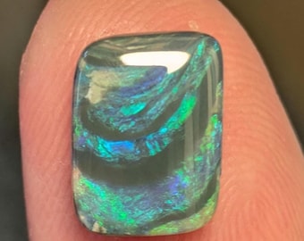 May include: A rectangular polished gemstone featuring a dark base color with vibrant, swirling iridescent patterns in shades of electric blue and bright green. The surface is smooth and reflective, displaying a natural play of color throughout.