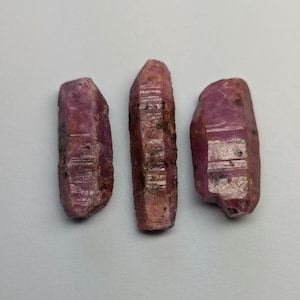 Op de afbeelding: Drie roze robijnkristallen, elk met een puntige punt en een ruw, getextureerd oppervlak.