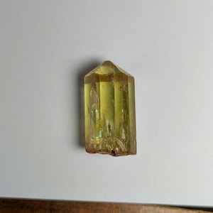 May include: A yellow crystal point with a pointed top and a flat base. The crystal is translucent and has a slight green hue.
