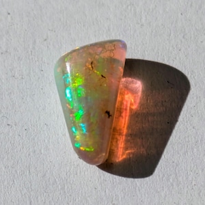 May include: A teardrop-shaped opal gemstone with a light pink, green, and iridescent sheen. The opal is set against a white background.