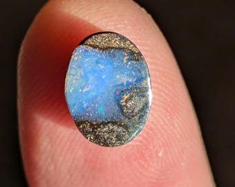 May include: A blue and black opal gemstone with a shimmering iridescent surface. The opal is oval shaped and has a dark, metallic base.