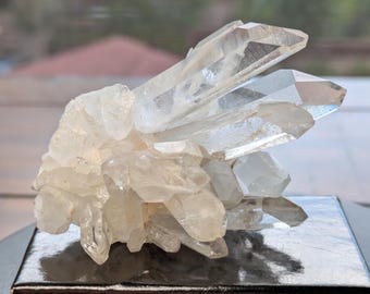 May include: A cluster of clear quartz crystals, with multiple points and a translucent appearance. The crystals are arranged in a natural, clustered formation, showcasing their natural beauty. The quartz sits on a reflective black surface.