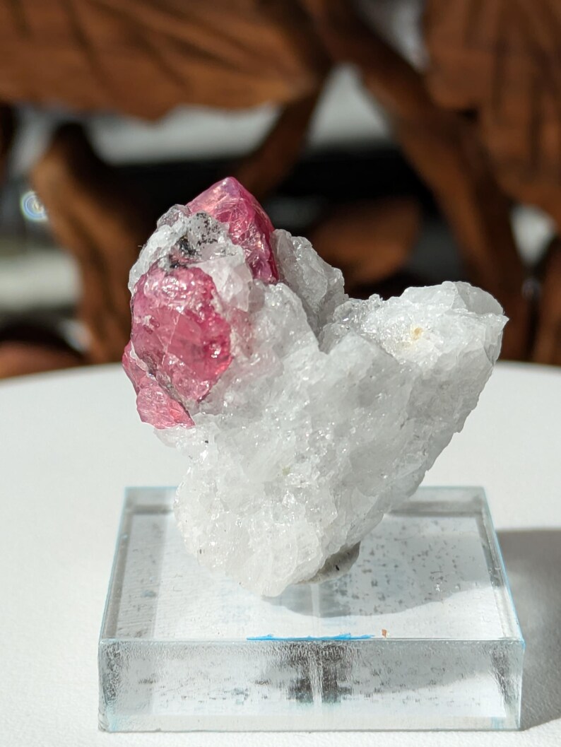 May include: A pink ruby crystal on a white matrix, displayed on a clear glass base.