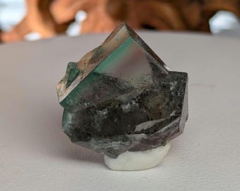 May include: A dark green, gray, and purple fluorite crystal with a white base. The crystal has a sharp, geometric shape.