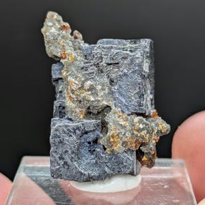 Könnte beinhalten: Ein metallisch graues Bleiglanz-Mineralexemplar mit kubischen Kristallstrukturen und einer rauen, texturierten Oberfläche. Eine Ansammlung kleiner, durchscheinender bernsteinfarbener Kristalle ist an der Seite der dunklen Formation eingebettet.