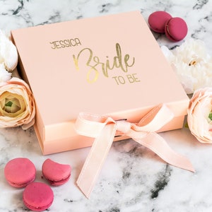 May include: A pink gift box with a gold ribbon and the text "Jessica Bride to Be" on the top. The box is surrounded by pink and white flowers and pink macarons.