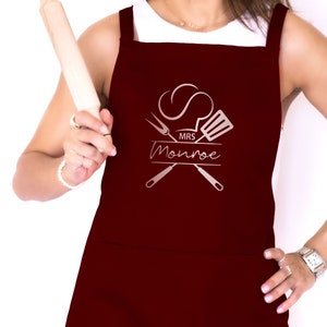 May include: Burgundy red apron with a white chef hat, crossed kitchen utensils, and the text "MRS Monroe" in a silver script font.