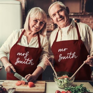 May include: Two people wearing matching burgundy aprons with the name "Smith" in white script. One apron says "mr" and the other says "mrs". They are in a kitchen, preparing food.