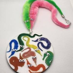 K&ouml;nnte beinhalten: Ein wei&szlig;er runder Filzbrett mit bunten Filz-Wurmformen und Glitzern. Zwei flauschige Wurmspielzeuge, eines rosa und eines gr&uuml;n, sind an einer silbernen Kette befestigt.