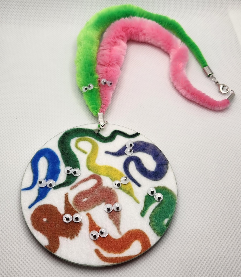 K&ouml;nnte beinhalten: Ein wei&szlig;er runder Filzbrett mit bunten Filz-Wurmformen und Glitzern. Zwei flauschige Wurmspielzeuge, eines rosa und eines gr&uuml;n, sind an einer silbernen Kette befestigt.