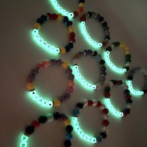 Peut inclure: Plusieurs bracelets de perles color&eacute;es avec des lettres blanches lumineuses &eacute;pelant des noms et des mots comme "Abstracted" et "Pomni". Les bracelets pr&eacute;sentent une vari&eacute;t&eacute; de couleurs de perles, dont le rouge, le bleu, le jaune et le violet, et sont illumin&eacute;s d'une lueur verte vive.