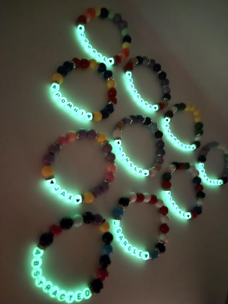 Peut inclure: Plusieurs bracelets de perles color&eacute;es avec des lettres blanches lumineuses &eacute;pelant des noms et des mots comme "Abstracted" et "Pomni". Les bracelets pr&eacute;sentent une vari&eacute;t&eacute; de couleurs de perles, dont le rouge, le bleu, le jaune et le violet, et sont illumin&eacute;s d'une lueur verte vive.