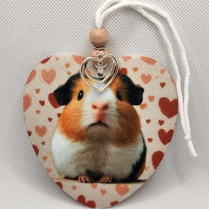 May include: A heart-shaped ornament featuring a cute guinea pig face and a pattern of pink and red hearts. The ornament has a brown wooden bead and a silver double heart charm attached to a white string.