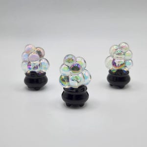 May include: Three decorative glass bubble sculptures. Each sculpture features a cluster of iridescent, spherical bubbles atop a small, black, cauldron-shaped base. The bubbles reflect a spectrum of colors, creating a shimmering effect.