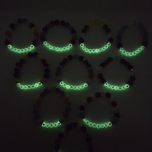 Peut inclure: Dix bracelets de perles avec des perles de lettres blanches lumineuses &eacute;pelant des noms tels que "Zooble", "Pragatha" et "Gummigoo". Les bracelets pr&eacute;sentent une vari&eacute;t&eacute; de perles color&eacute;es, dont le rouge, le noir et le jaune, sur fond sombre.