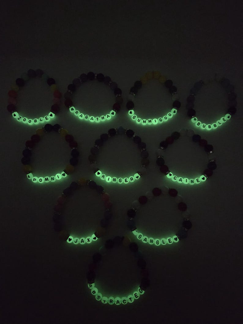 Peut inclure: Dix bracelets de perles avec des perles de lettres blanches lumineuses &eacute;pelant des noms tels que "Zooble", "Pragatha" et "Gummigoo". Les bracelets pr&eacute;sentent une vari&eacute;t&eacute; de perles color&eacute;es, dont le rouge, le noir et le jaune, sur fond sombre.