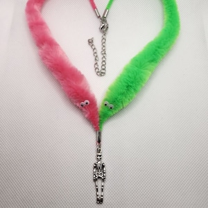 Puede incluir: Un collar de gusano peludo rosa y verde con ojos saltones y un dije de esqueleto plateado.