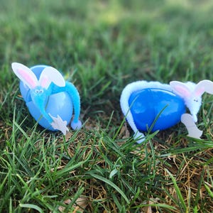 May include: Two blue Easter egg decorations with white and pink bunny features are set in green grass. One bunny has blue arms and the other has white fur around the egg. The bunny ears are pink and white.