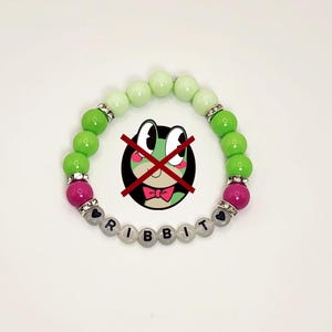 May include: A beaded bracelet with green, pink, and clear beads. The bracelet spells out the word "RIBBIT" with letter beads. The beads are strung on a clear elastic cord. The bracelet has silver-colored spacers.