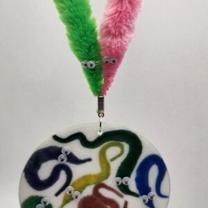K&ouml;nnte beinhalten: Eine runde, wei&szlig;e, Plastikplatte mit einem bunten Design aus regenbogenfarbenen W&uuml;rmern mit Glitz Augen. Zwei flauschige, bunte wurmf&ouml;rmige Spielzeuge sind mit einem silbernen Clip an der Oberseite der Platte befestigt.