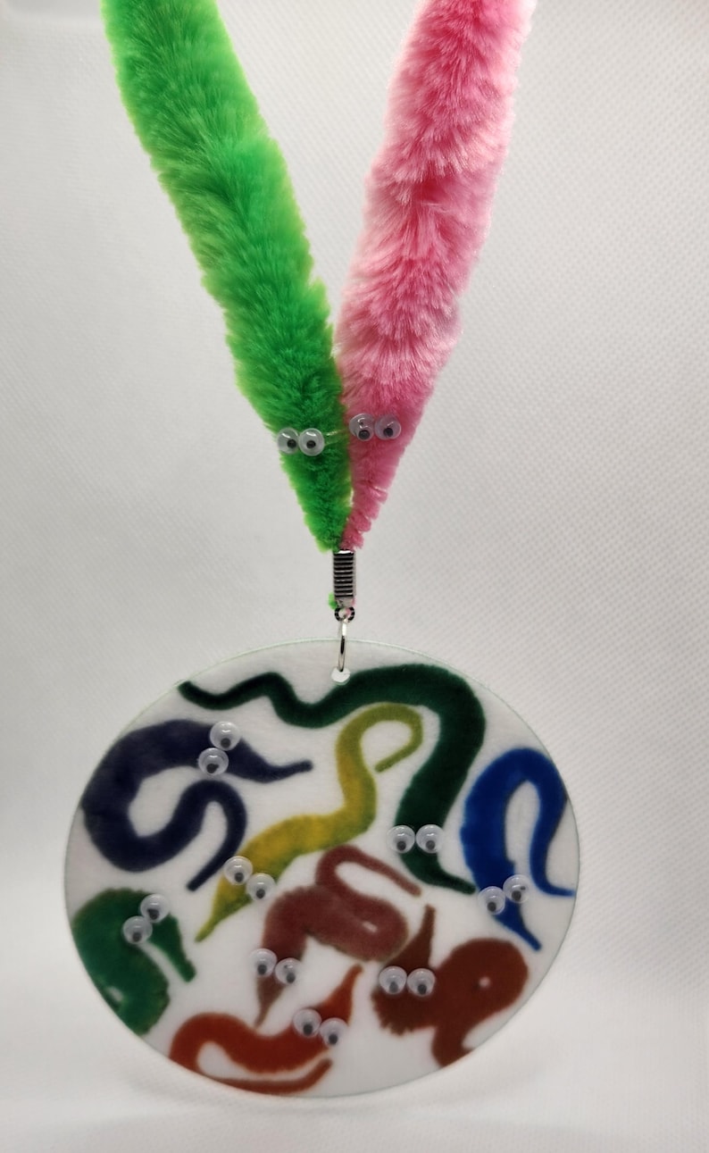 K&ouml;nnte beinhalten: Eine runde, wei&szlig;e, Plastikplatte mit einem bunten Design aus regenbogenfarbenen W&uuml;rmern mit Glitz Augen. Zwei flauschige, bunte wurmf&ouml;rmige Spielzeuge sind mit einem silbernen Clip an der Oberseite der Platte befestigt.
