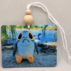 Puede incluir: Un juguete de peluche azul y amarillo de un personaje de Star Wars sentado en una playa de arena con un cuerpo de agua en el fondo. El juguete está unido a una cuenta de madera y una cuerda blanca.