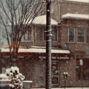 Könnte beinhalten: Winterliche Straßenszene mit einem beleuchteten, verzierten Laternenpfahl im Vordergrund. Im Hintergrund sind Gebäude und kahle Bäume zu sehen. Das Schild "Villar Deli & Catering" ist erkennbar. Schnee bedeckt die Szene.