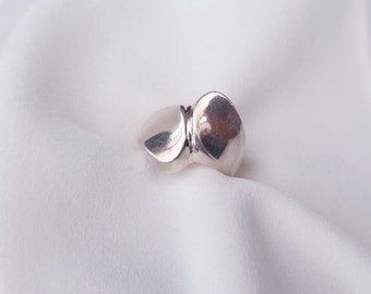 Könnte beinhalten: Ein silberner Ring mit einem einzigartigen, abstrakten Design. Der Ring hat zwei abgerundete, unregelmäßige Formen, die durch ein schmales Band verbunden sind. Das polierte Metall reflektiert das Licht und erzeugt Glanzlichter und Schatten. Der Ring ist vor weißem Hintergrund platziert.