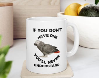 May include: White ceramic mug featuring a gray parrot with a red tail. The mug has the text "IF YOU DON'T HAVE ONE YOU'LL NEVER UNDERSTAND" in black. The mug is sitting on a beige coaster.