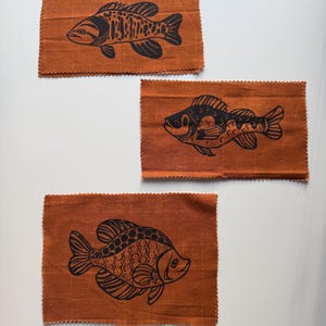 May include: Three orange fabric patches with black fish illustrations. Each patch features a different fish design, with detailed linework. The fabric has a textured appearance and a scalloped edge. The fish designs vary in style and detail.