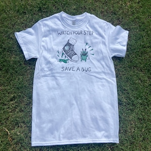 May include: White t-shirt with a graphic design featuring a sneaker stepping on a bug, with the text "WATCH YOUR STEP" above and "SAVE A BUG" below. The design is in black, green, and white.