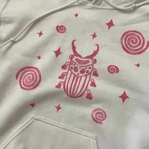 K&ouml;nnte beinhalten: Wei&szlig;er Kapuzenpullover mit einem rosa K&auml;fer-Motiv, Spiralen und Sternformen. Der Pullover hat eine Vordertasche und eine Kapuze mit Kordelzug. Das K&auml;fer-Design ist mittig auf der Vorderseite.