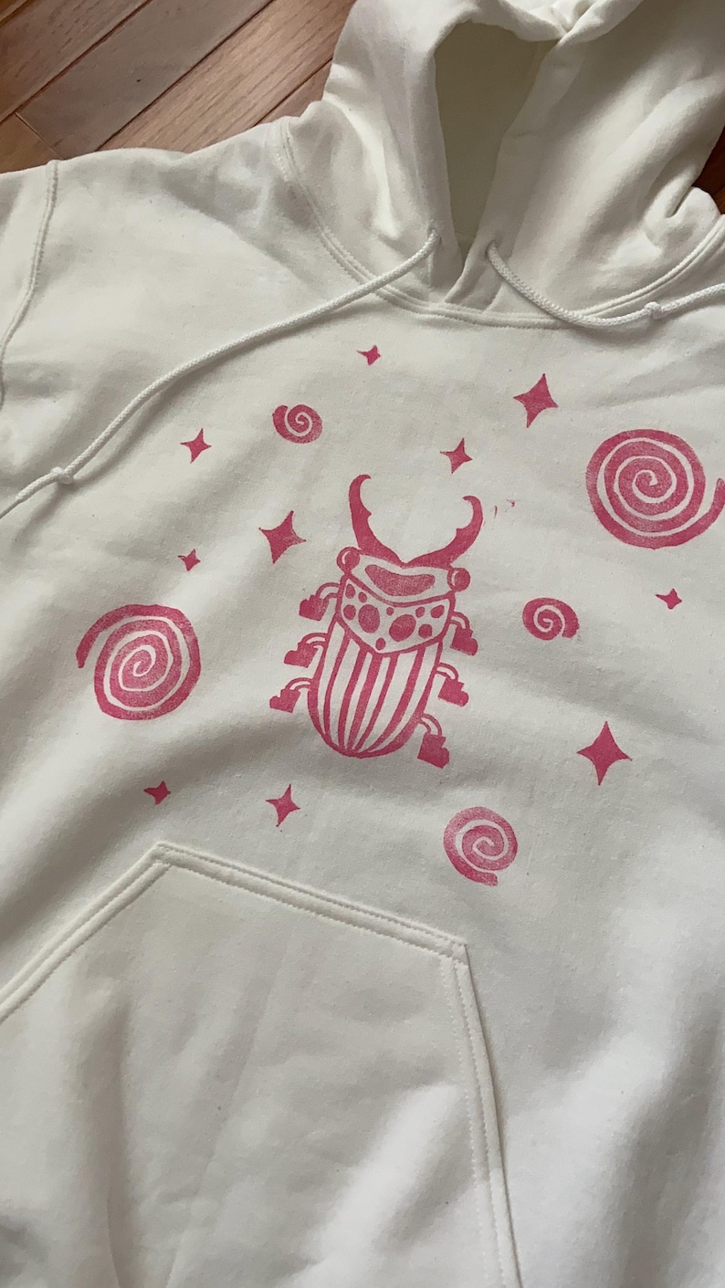 K&ouml;nnte beinhalten: Wei&szlig;er Kapuzenpullover mit einem rosa K&auml;fer-Motiv, Spiralen und Sternformen. Der Pullover hat eine Vordertasche und eine Kapuze mit Kordelzug. Das K&auml;fer-Design ist mittig auf der Vorderseite.