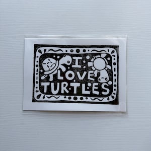 May include: Black and white illustration on a white background, featuring the text "I LOVE TURTLES" with a turtle and a sun graphic. The design is enclosed in a dotted border.