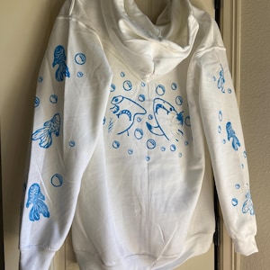 May include: White hooded sweatshirt with blue goldfish and bubble graphics. The back features two large goldfish and bubbles, while the sleeves display smaller goldfish. The design is printed in a vibrant blue color.