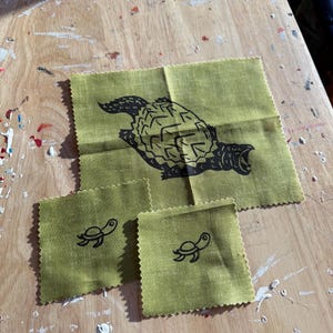May include: A set of four olive green fabric squares with black turtle designs. The largest square features a detailed turtle illustration, while the smaller squares have simpler turtle outlines. The fabric has a textured appearance.