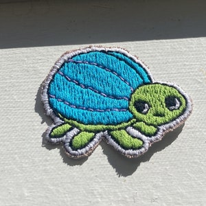 May include: Embroidered turtle patch featuring a blue shell with purple stitching and a green head and limbs. The patch has a white border and is designed to be sewn onto fabric. The turtle has a friendly expression.