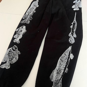 May include: Black sweatpants featuring a repeating pattern of detailed white fish illustrations. The fish vary in size and species, creating a unique and artistic design. The pants are made of a soft material.