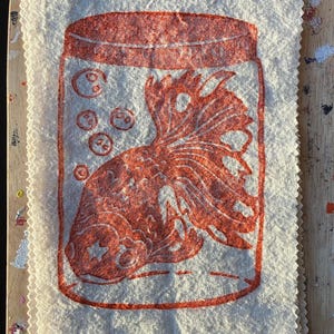 May include: A cream-colored fabric square with a red print of a goldfish in a jar. The jar has bubbles and a lid. The goldfish has detailed scales and fins. The fabric has a textured appearance and a serged edge.