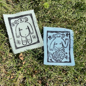 May include: Two blue fabric patches with black embroidered designs. One patch features a cartoon-like figure with a large head and a smile. The other patch features a mermaid with a tail and a star design.