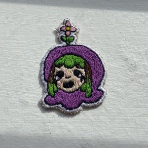 May include: Embroidered patch featuring a cartoon character with green hair and a purple hood. The character has a sad expression and is topped with a small flower. The patch has a white border and is likely for clothing or accessories.