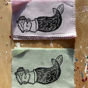 May include: Two fabric squares with a black and white illustration of a mermaid holding a book. The top square is pink and the bottom square is green.