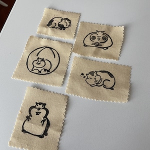 May include: Five small, beige fabric patches with black ink illustrations of hamsters in various poses. One hamster is curled up, another is in a ball, and others are in different positions. The patches have a zig-zag edge.