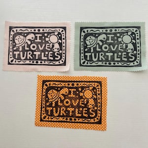 May include: Three fabric patches with the text "I LOVE TURTLES" and turtle illustrations. One patch is light pink, another is light green, and the third is orange with white polka dots. The text and turtle designs are in black.