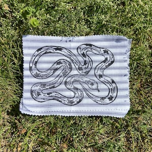 May include: A white fabric square with a black and white snake design printed on it. The fabric has a striped pattern.