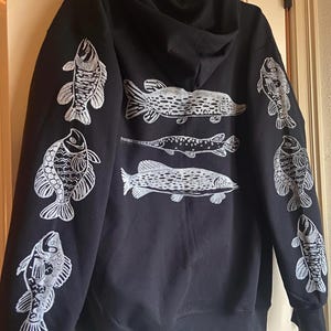 May include: Black hooded sweatshirt with a white print of various fish on the front and sleeves. The fish are stylized and have a vintage look.