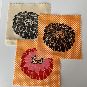 May include: Three fabric swatches with floral designs. One swatch is beige with a black flower print. Two swatches are orange with white polka dots, one with a brown flower print and the other with a pink and red flower print.