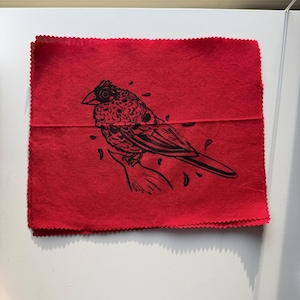 May include: A red square cloth with a black illustration of a bird perched on a branch. The bird has detailed feathers and is surrounded by small leaf-like shapes. The edges of the cloth have a decorative, zig-zag pattern.