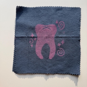 May include: A navy blue square cloth with a pink tooth graphic. The tooth has a grumpy expression and is surrounded by swirls and stars. The edges of the cloth are scalloped.